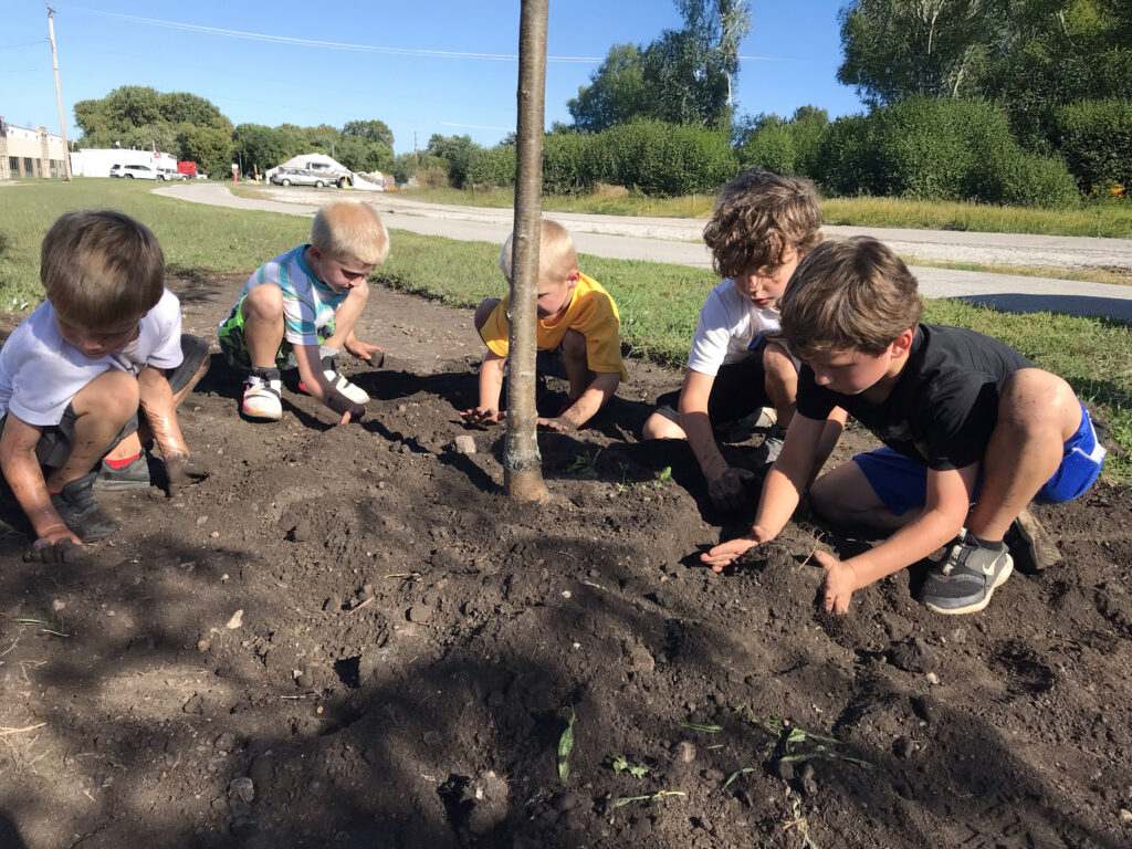 Five young children kneeling in the soil around a newly planted tree, patting the earth into place.
