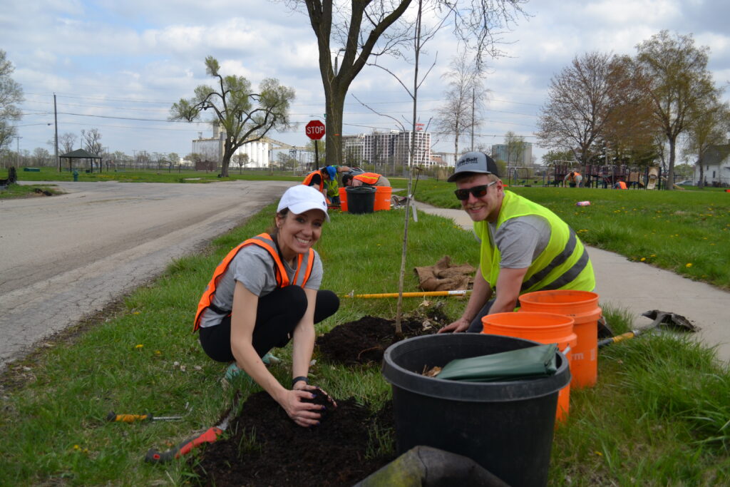 Two volunteers in safety vests kneeling beside a newly planted street tree, surrounded by buckets and tools.