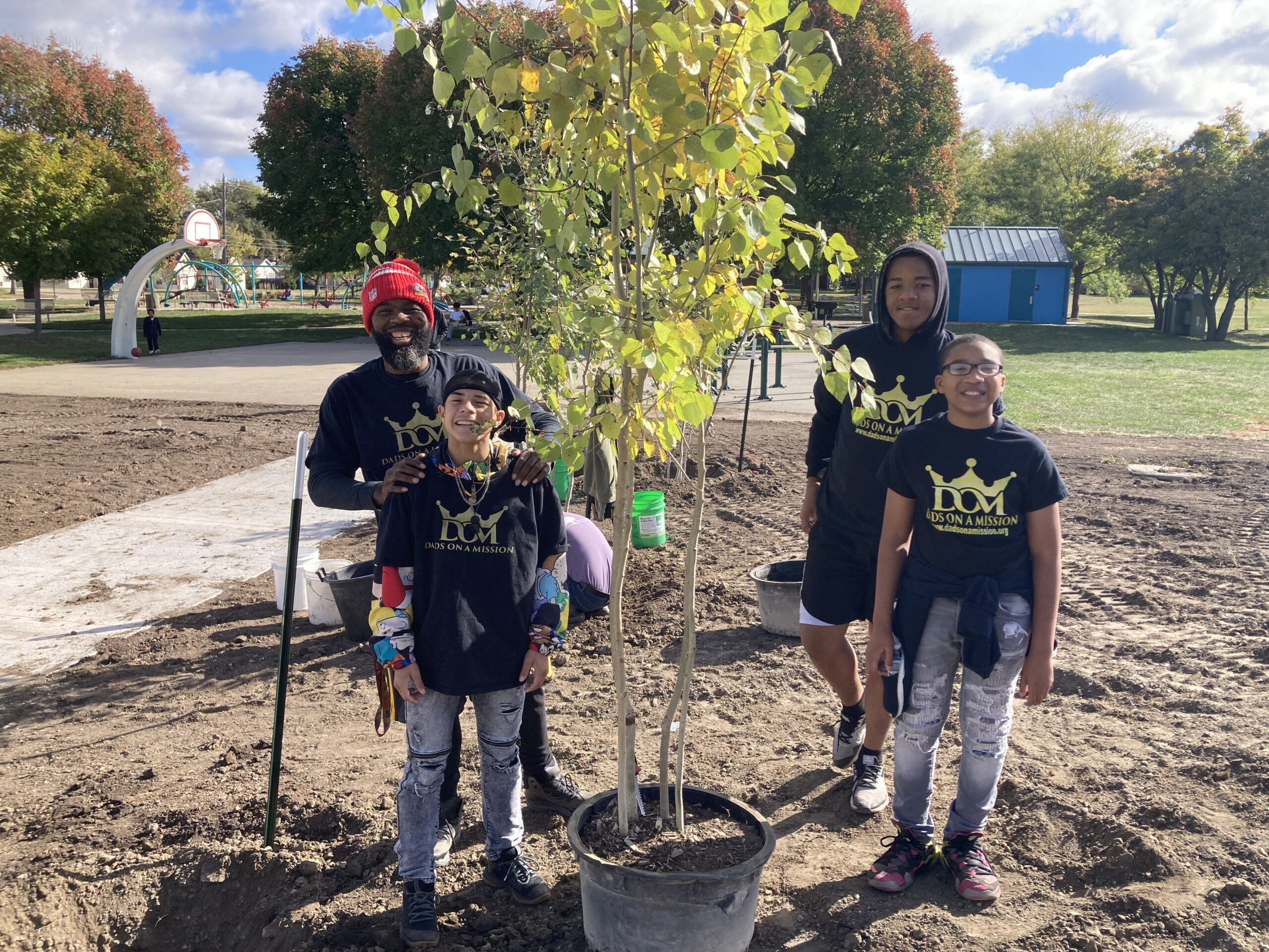Volunteers in "Kids on a Mission" shirts standing beside a young tree ready for planting in a city park.