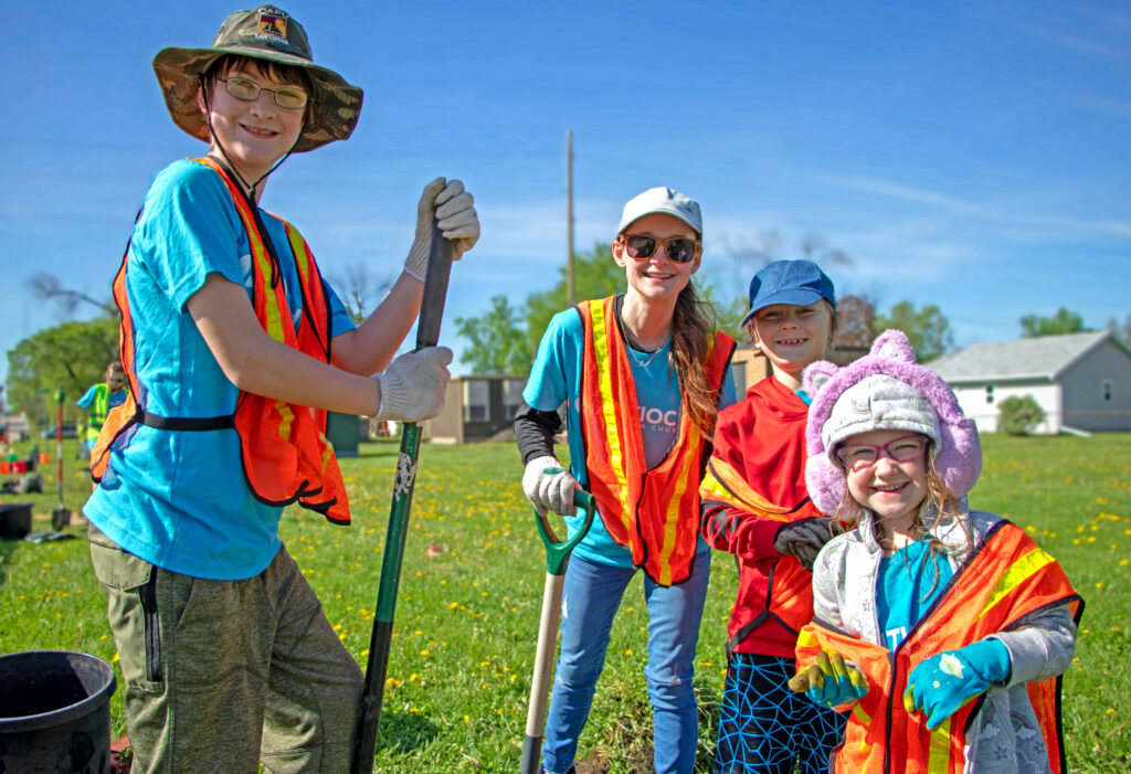 Four young volunteers in safety vests holding shovels on a grassy planting site.