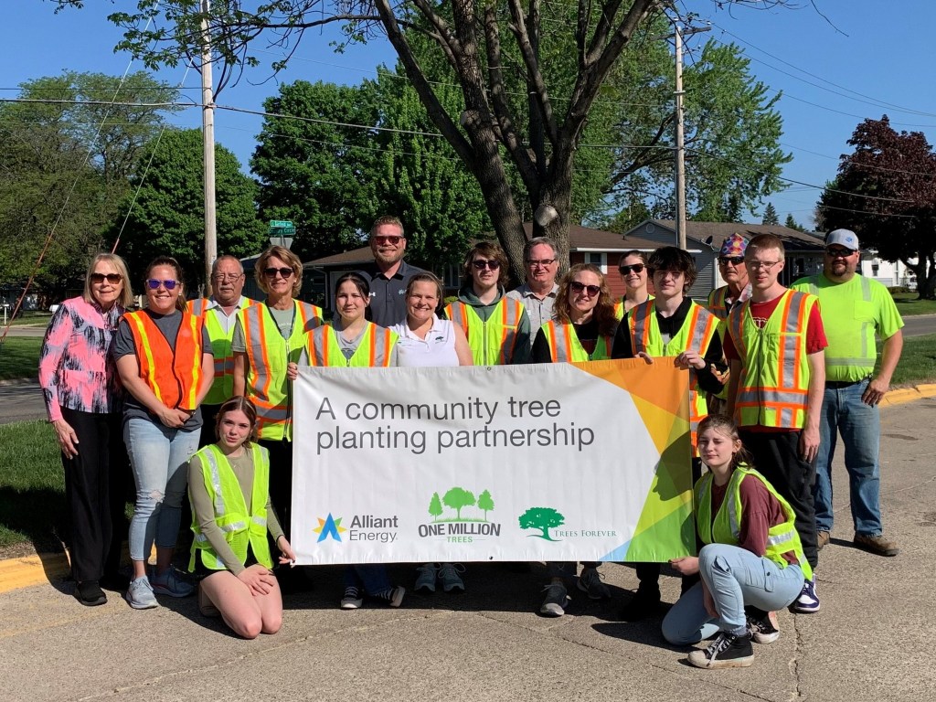 A large group of volunteers in high-visibility vests holding a banner reading "A community tree planting partnership".
